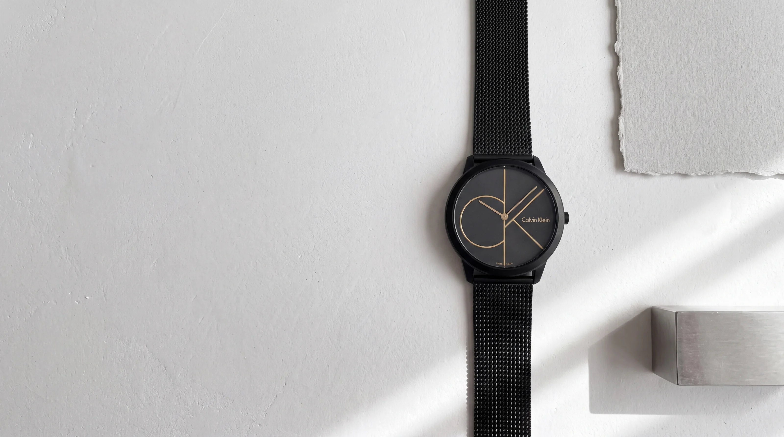 Calvin Klein Minimal K3M214X1 black watch with a large gold ck monogram dial, on white plaster in window light