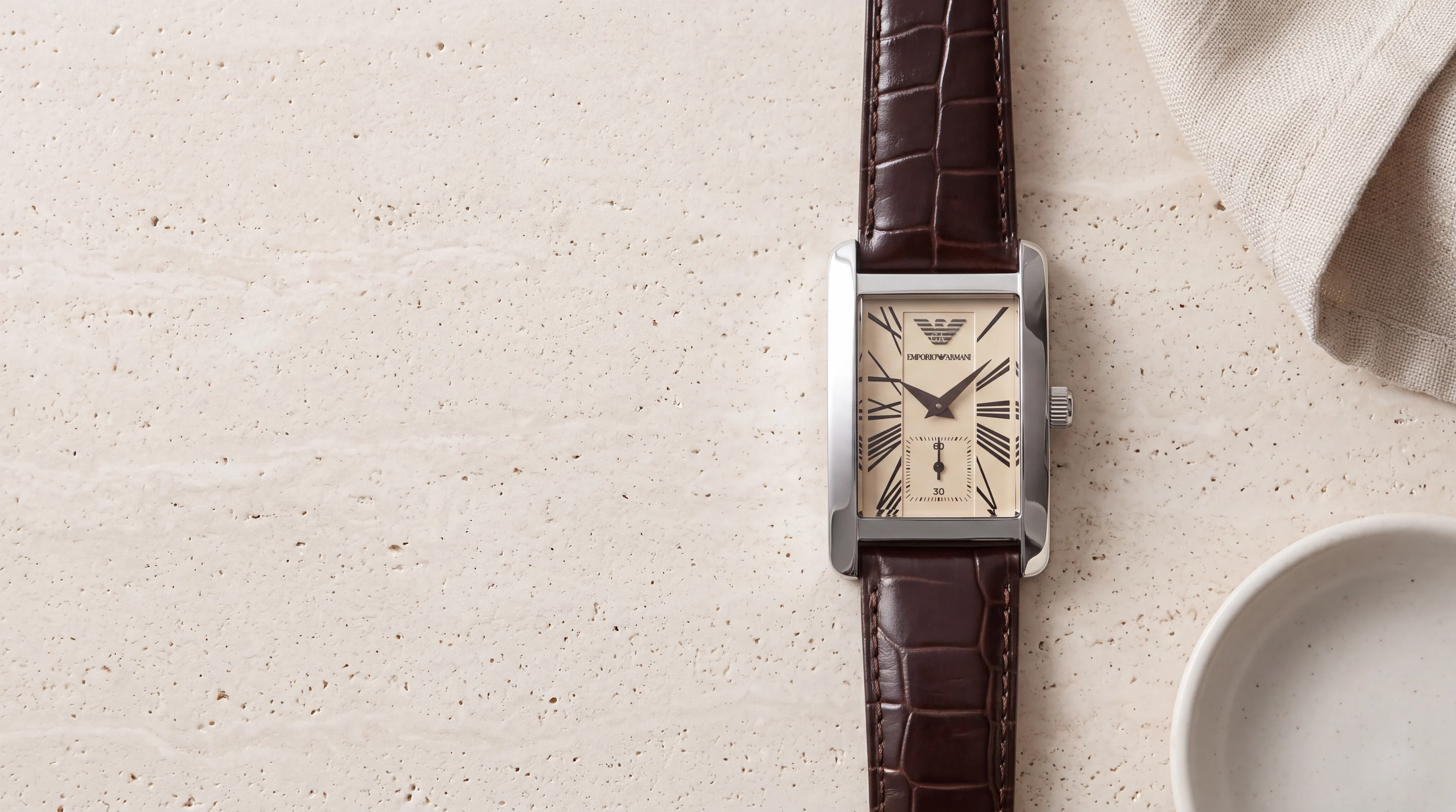 Montre Emporio Armani AR0154 rectangulaire en acier, cadran crème à chiffres romains, cuir marron, sur travertin