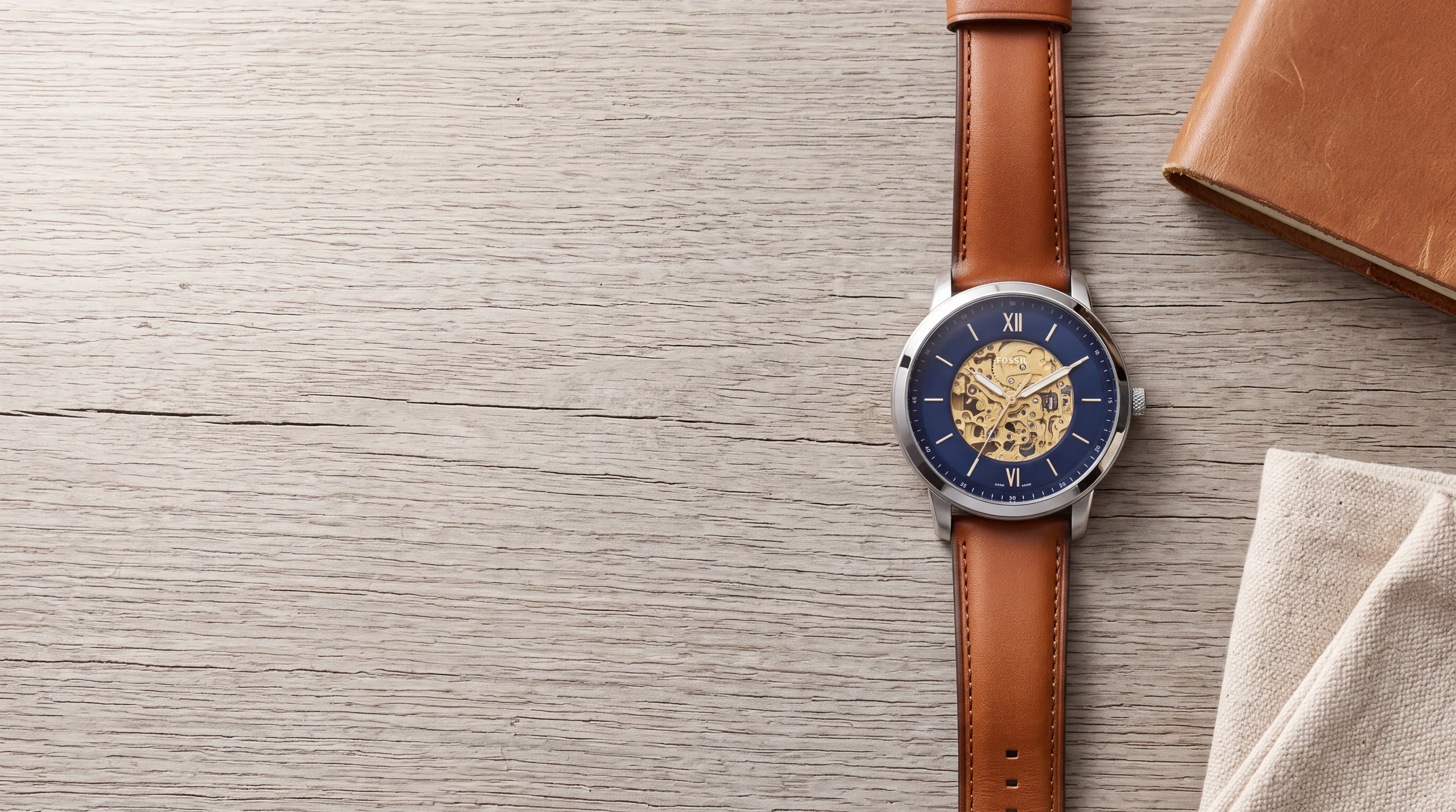 Fossil Neutra Automatic ME3160 with open-worked blue dial on raw oak beside a leather road-trip notebook