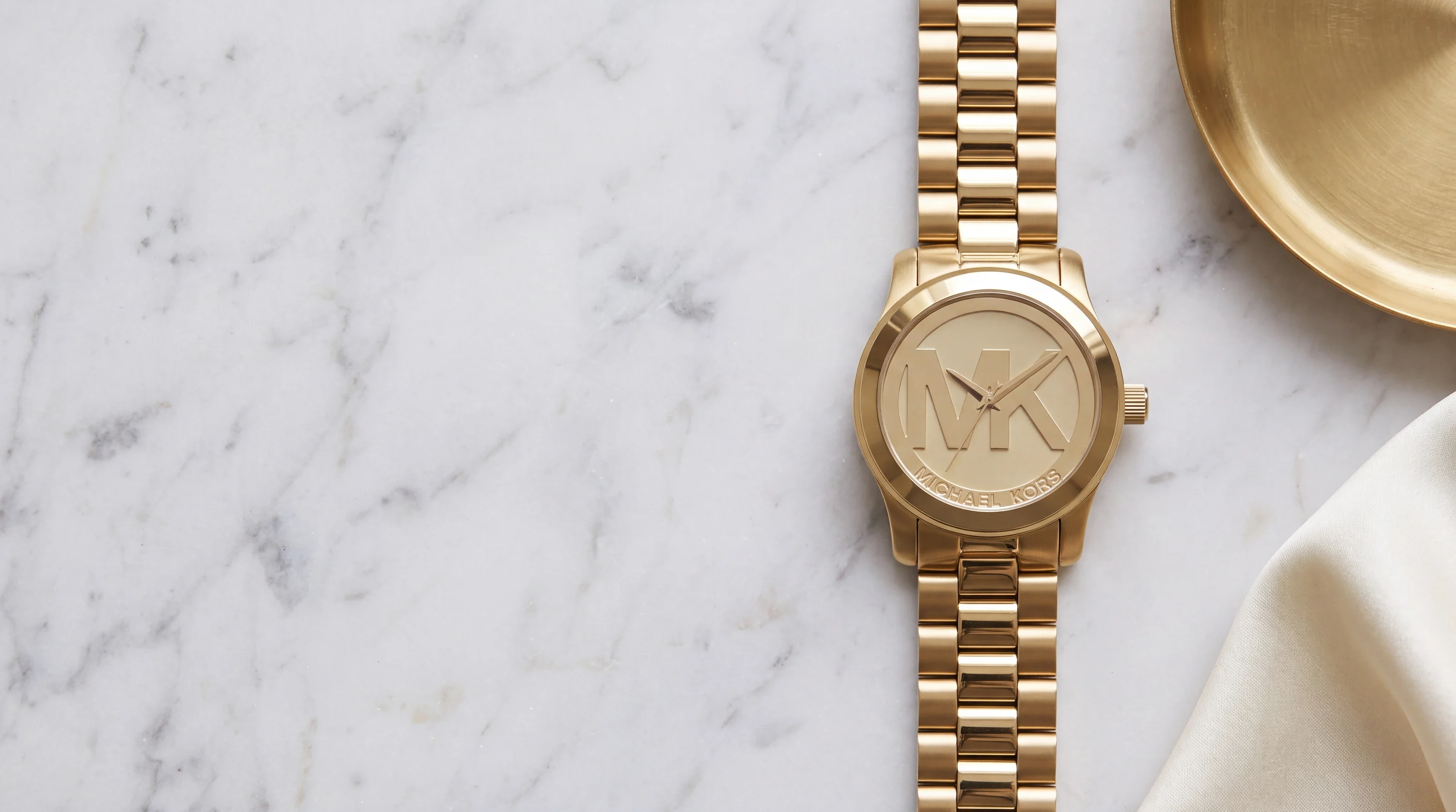 Michael Kors Runway MK5473 gold-tone watch with large MK logo dial on Carrara marble beside a brass tray