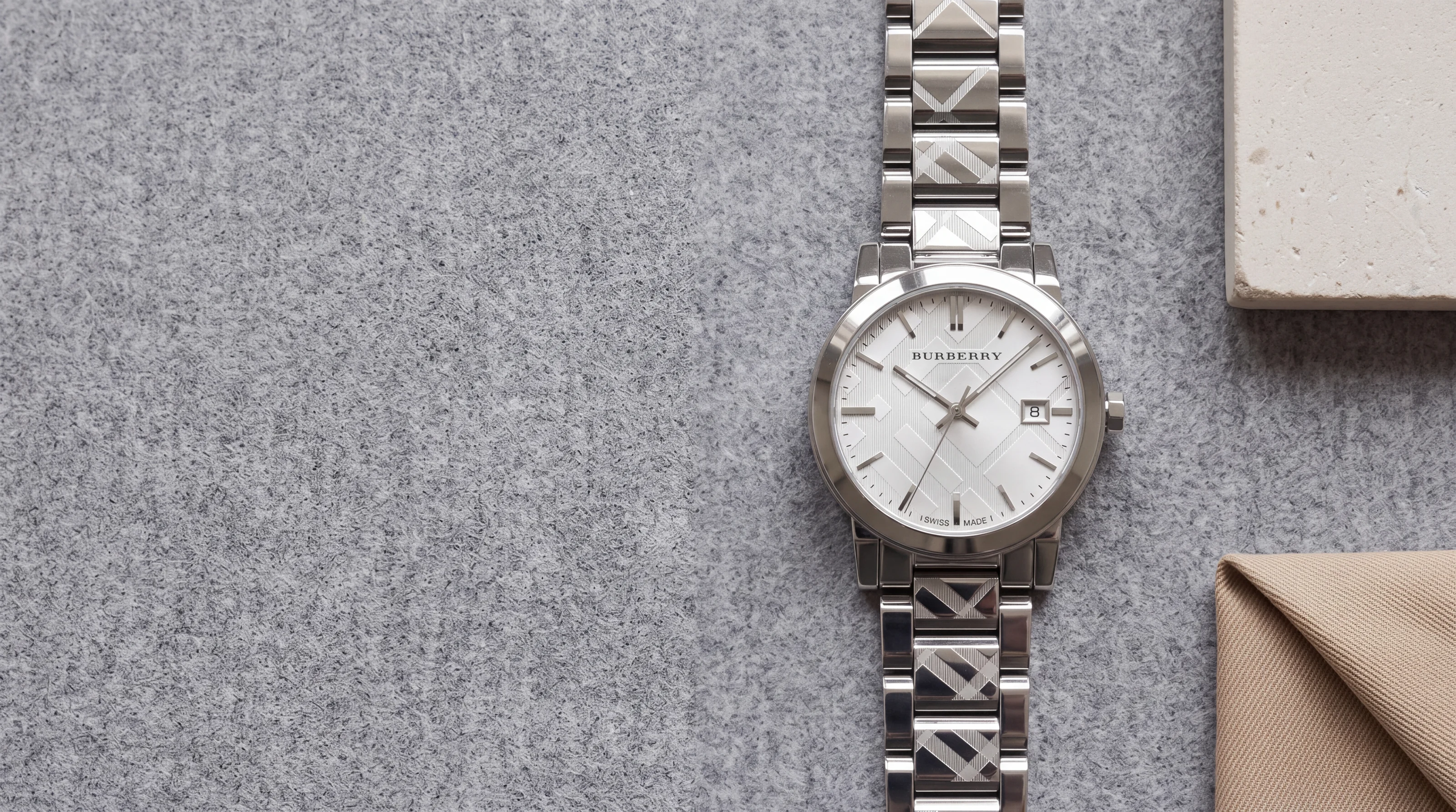 Montre Burberry The City BU9037 en acier, maillons gravés du motif check, posée sur une flanelle de laine grise