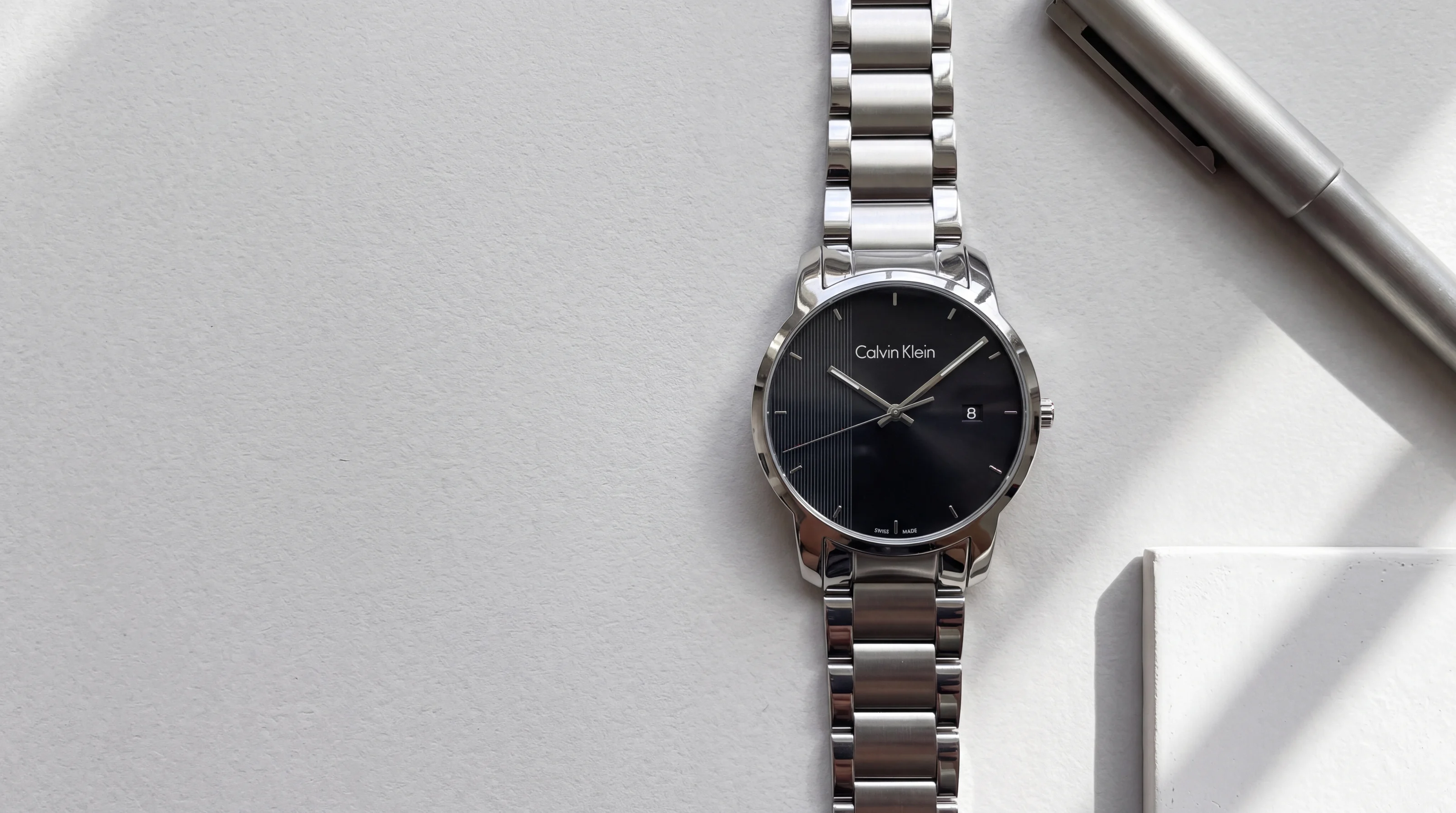 Montre homme Calvin Klein City K2G2G14Y en acier, cadran noir à panneau rayé, sur papier gris perle près d'un stylo