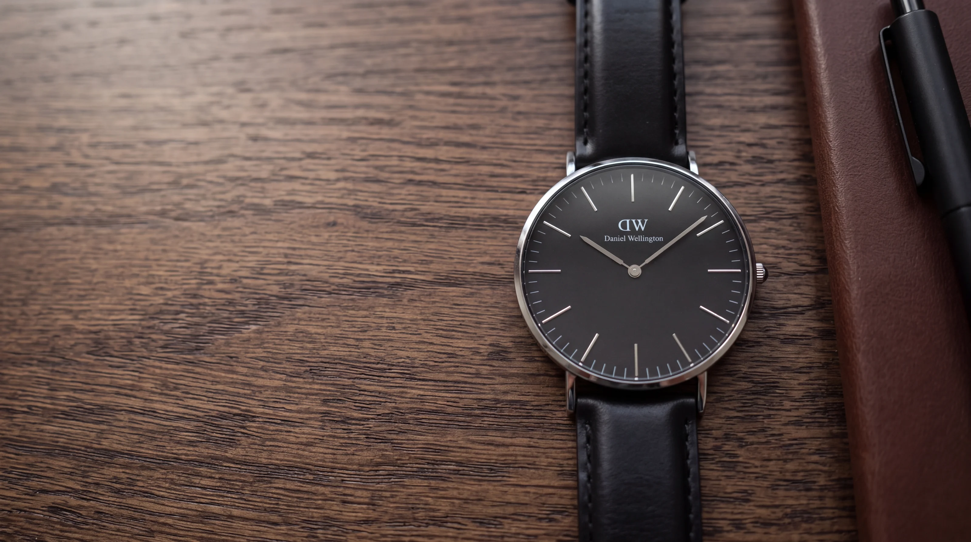 Montre Daniel Wellington Classic Black 40 mm, cadran noir et cuir noir, sur un bureau en chêne foncé avec un carnet
