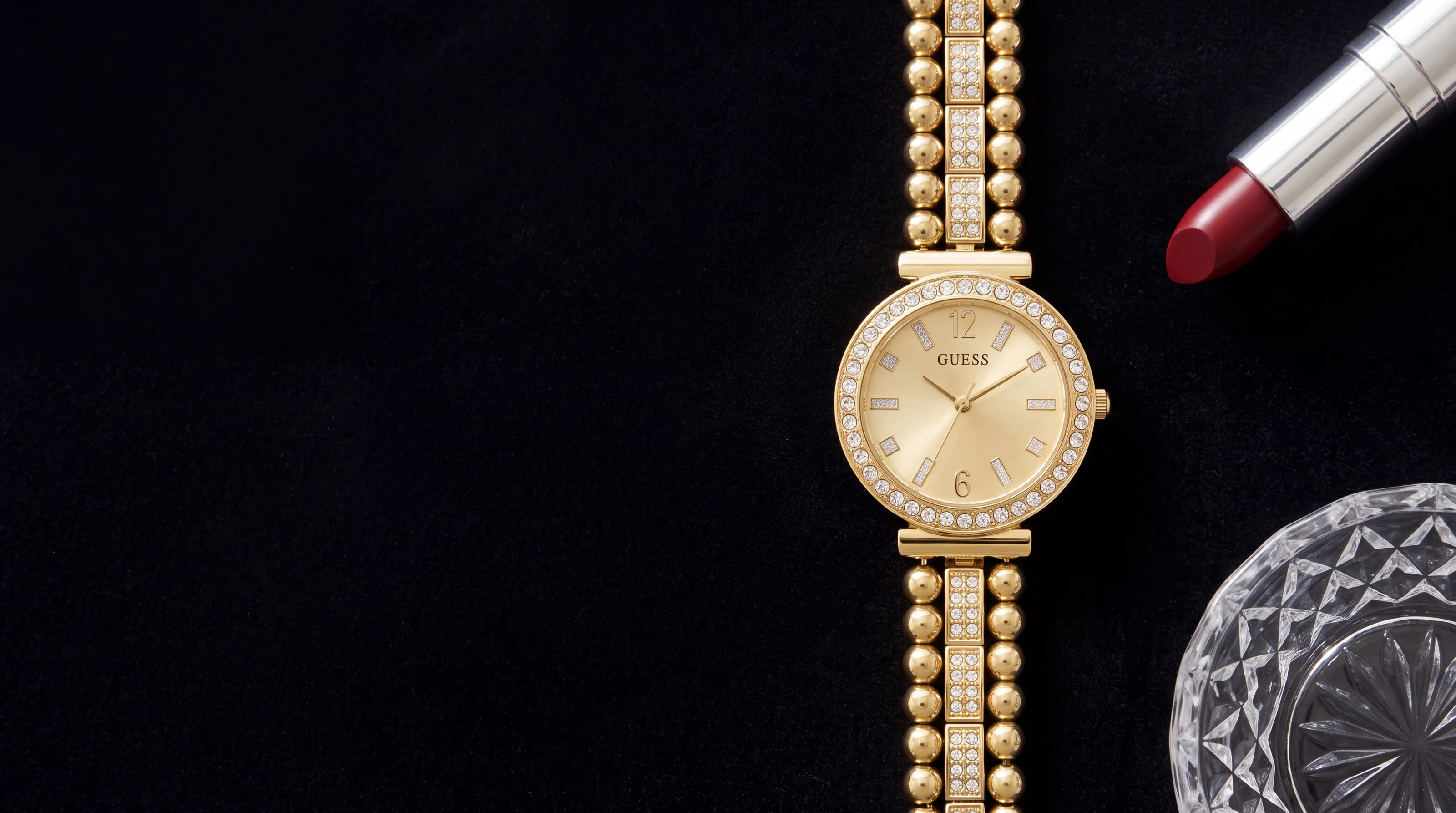 Montre femme GUESS Gala GW0401L2 dorée, lunette sertie de cristaux, sur velours noir près d'un rouge à lèvres