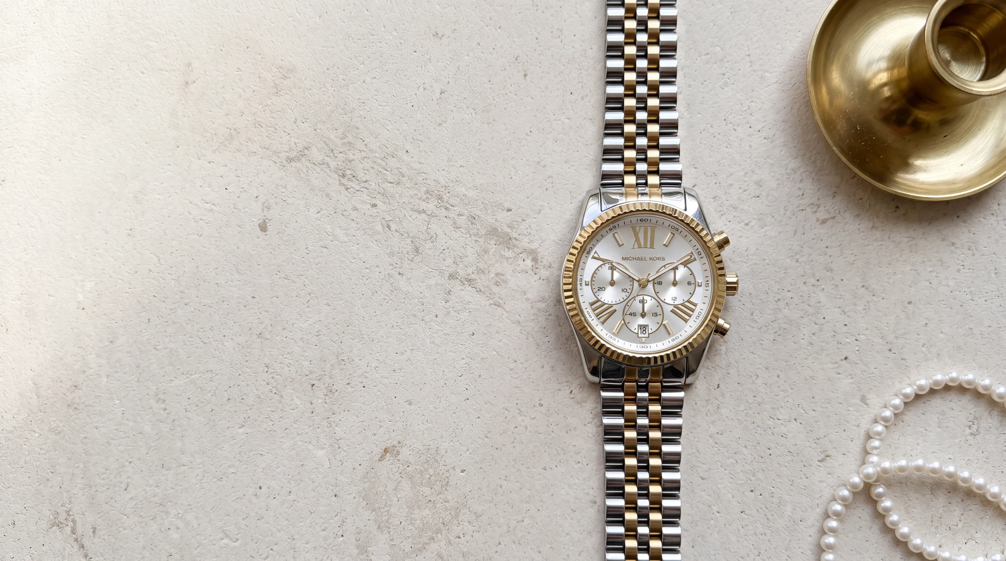 Montre Michael Kors Lexington MK5955 bicolore acier et or, chronographe posé sur pierre calcaire près de perles